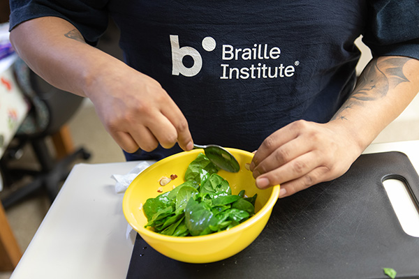 A man's hands stir a green spinach salad in a yellow bowl sitting on a black cutting board. 