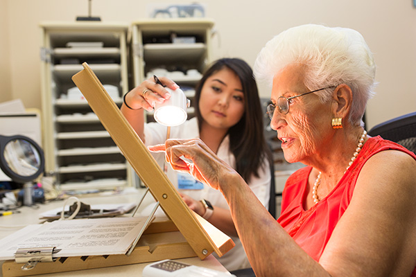 Senior woman reads words on an easel highlighted by task lighting while an instructor adjusts the light. 