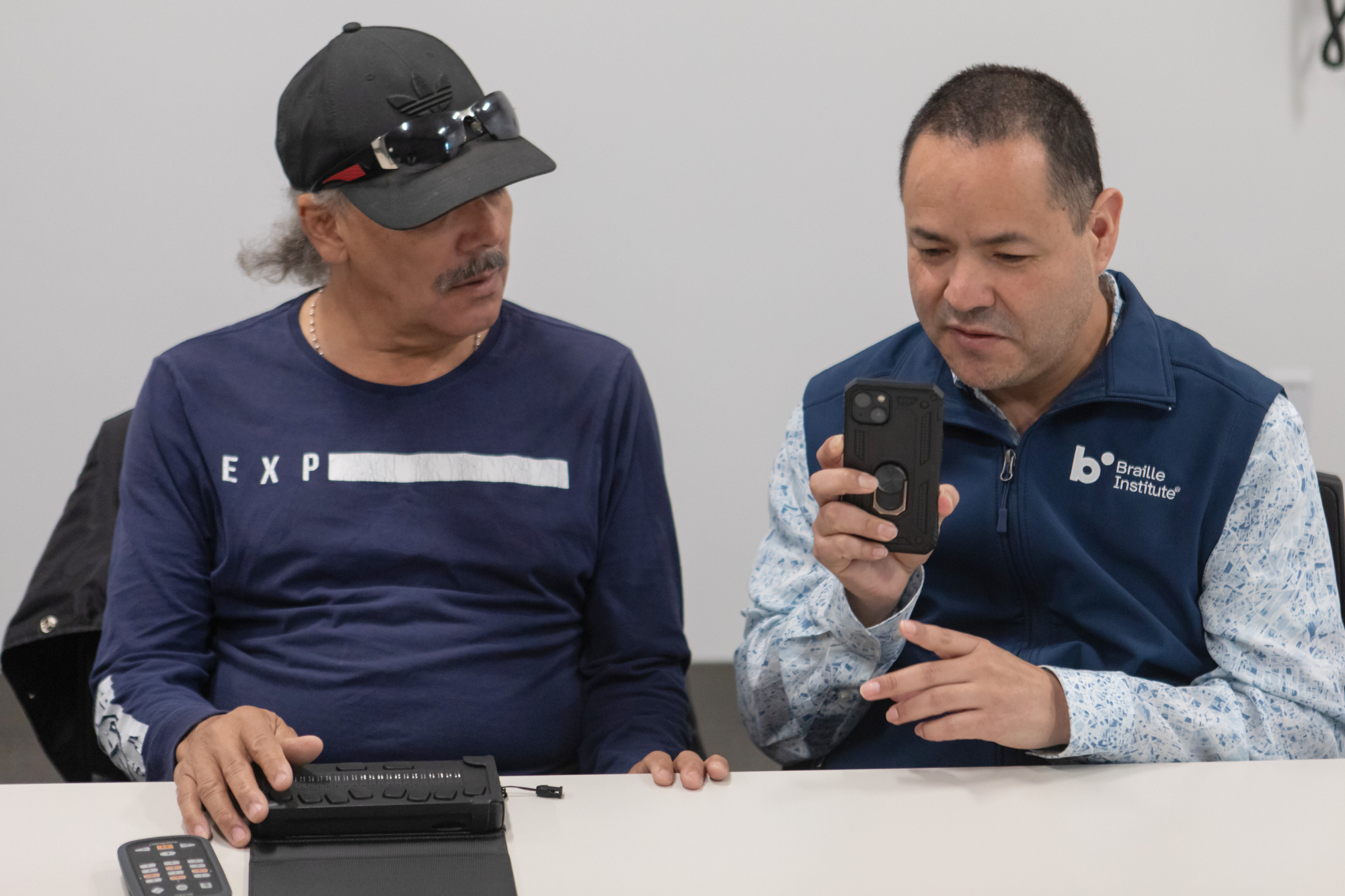 A Braille Institute student and Technology Instructor sit together at a table with several devices. The student has a braille e-reader and a Victor Reader in front of him while the instructor holds an iPhone up for a demonstration.