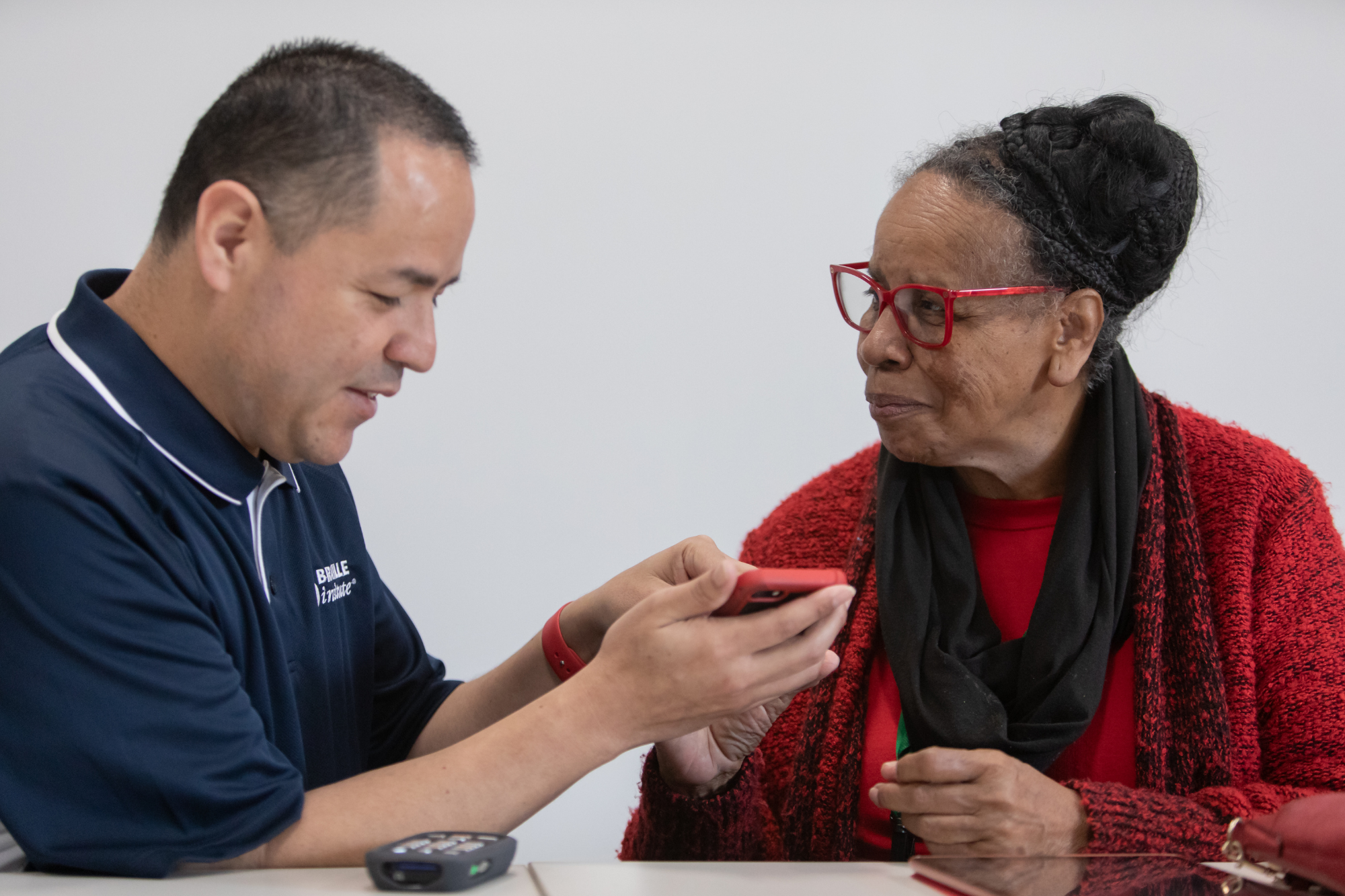 A Braille Institute Access Technology instructor sits with a student, showing her how to use VoiceOver on a phone in a red case. The instructor wears a blue Braille Institute polo shirt, and the student wears a red sweater and red glasses.