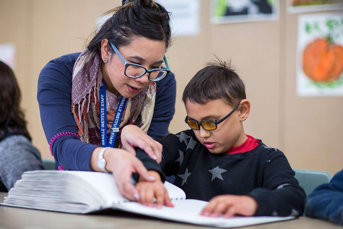 Mujer con gafas sujetando el dedo de un niño pequeño mientras le ayuda a pasarlo por un libro en braille.