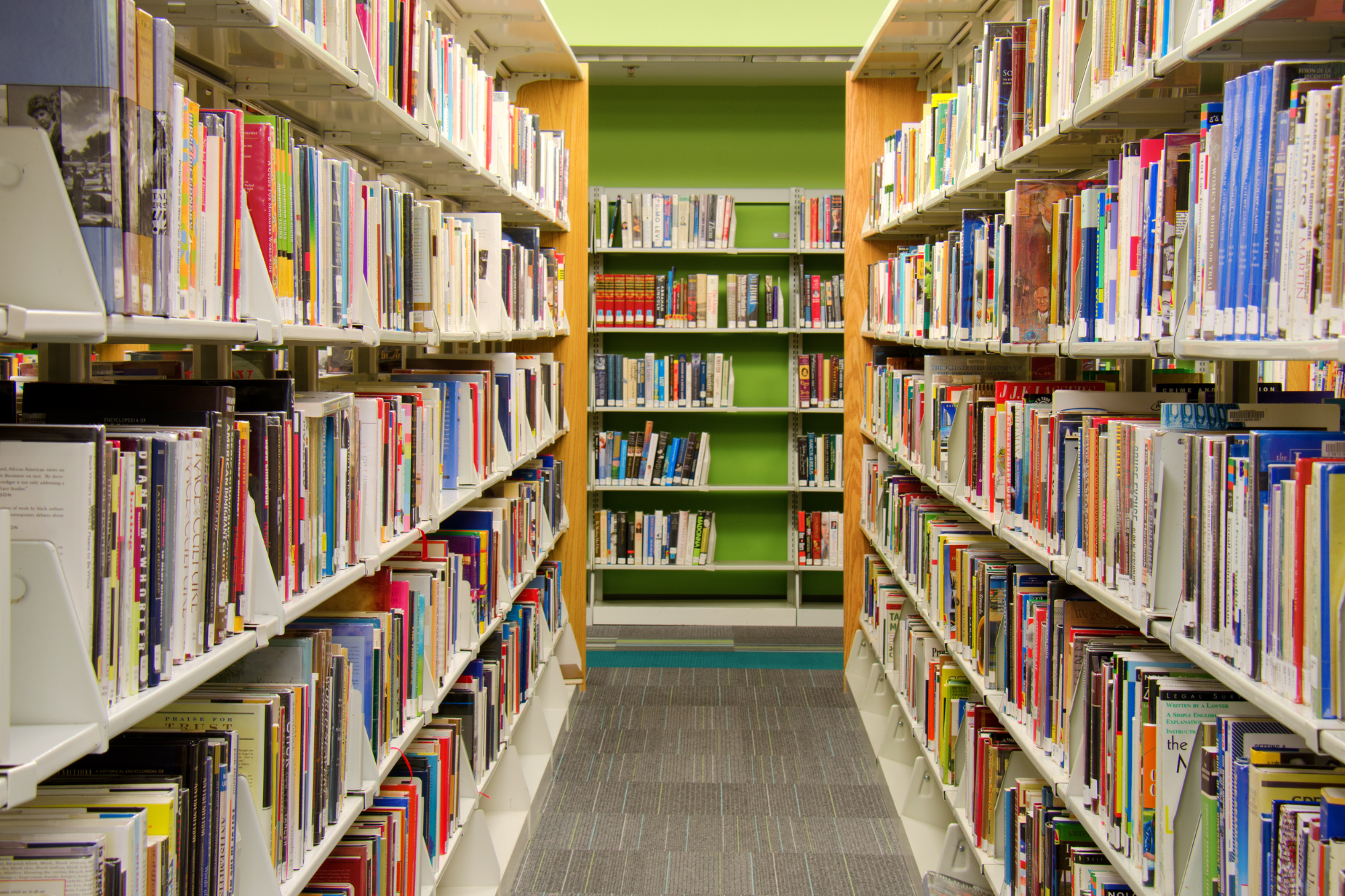 A view between two large, full bookshelves in a public library, facing another set of shelves against a green wall.