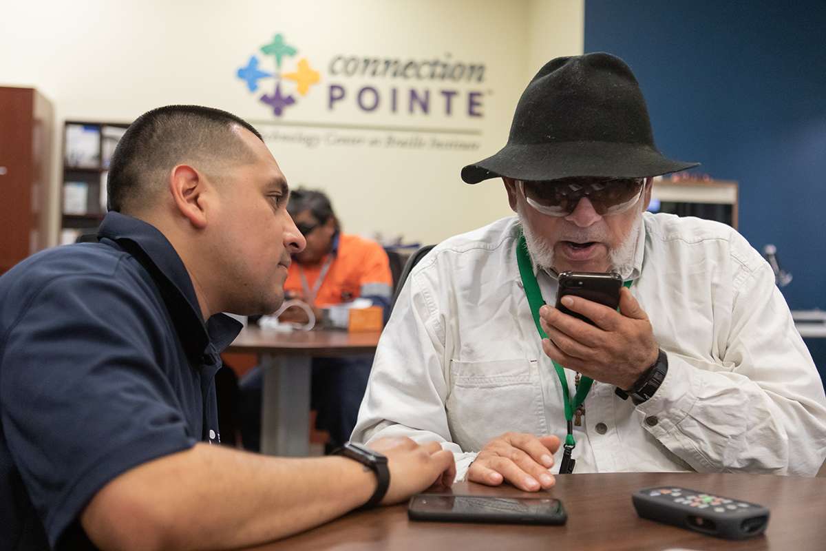 A man in a black wide brimmed hat speaks into a small device while a braille employee looks on. Both seated at a desk.
