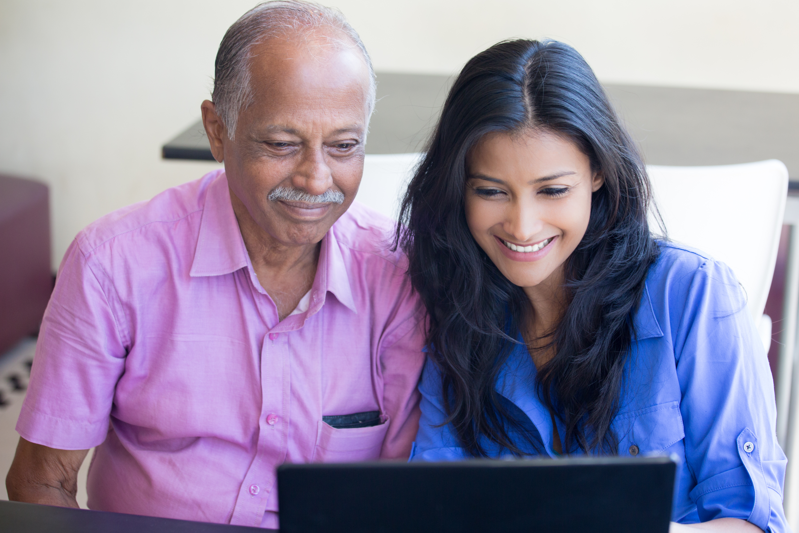 A Hispanic woman smiles as she shows her elderly father something on an open laptop.