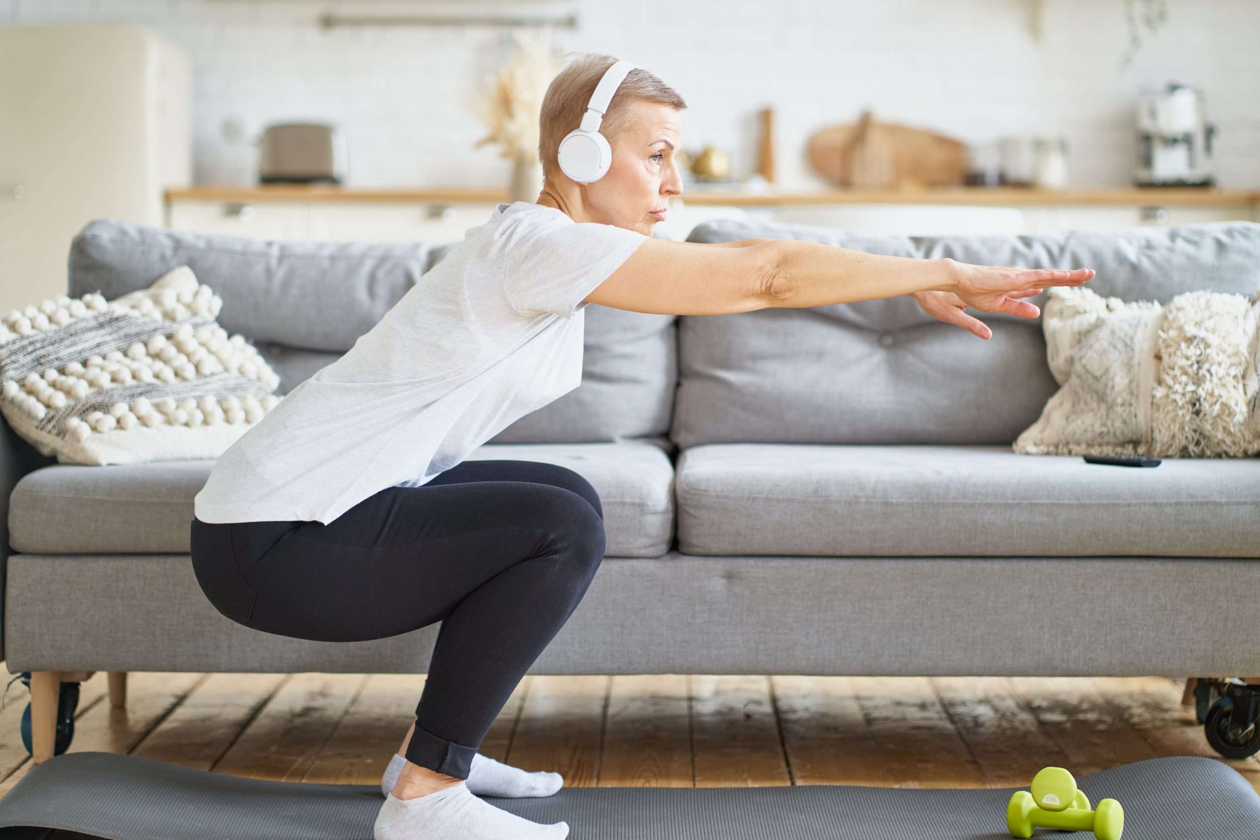 Una señora mayor con auriculares está sobre una colchoneta de ejercicios haciendo sentadillas con los brazos extendidos para mantener el equilibrio.
