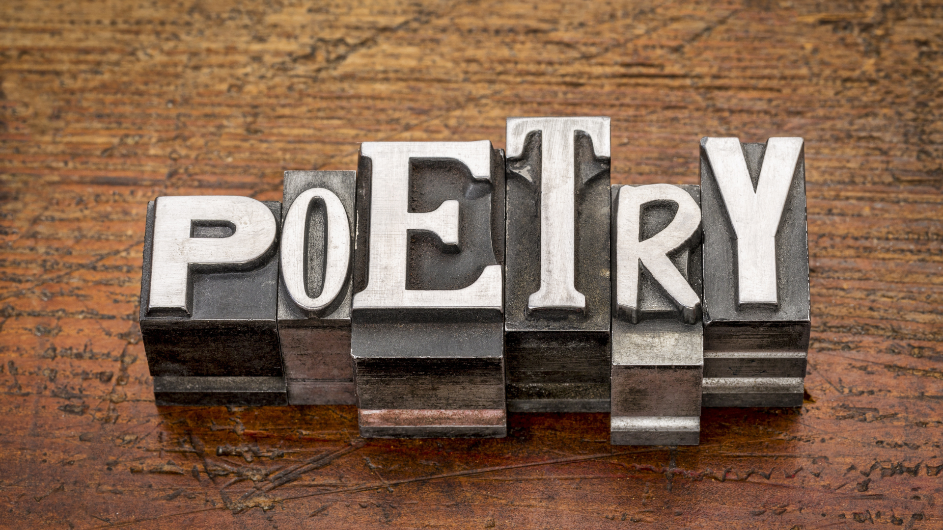 Mixed vintage metal type printing blocks spelling out the word "poetry" atop a wood surface.