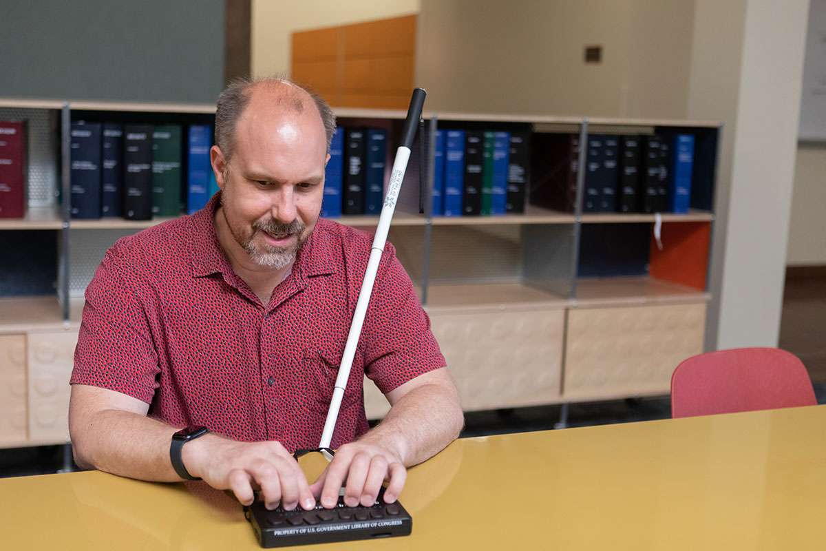 A man in a red shirt sits at a desk with a white cane next to him and hands on a brailler
