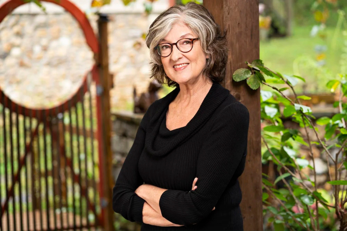 Barbara Kingsolver headshot. She smiles outdoors, leaning against a wooden post.