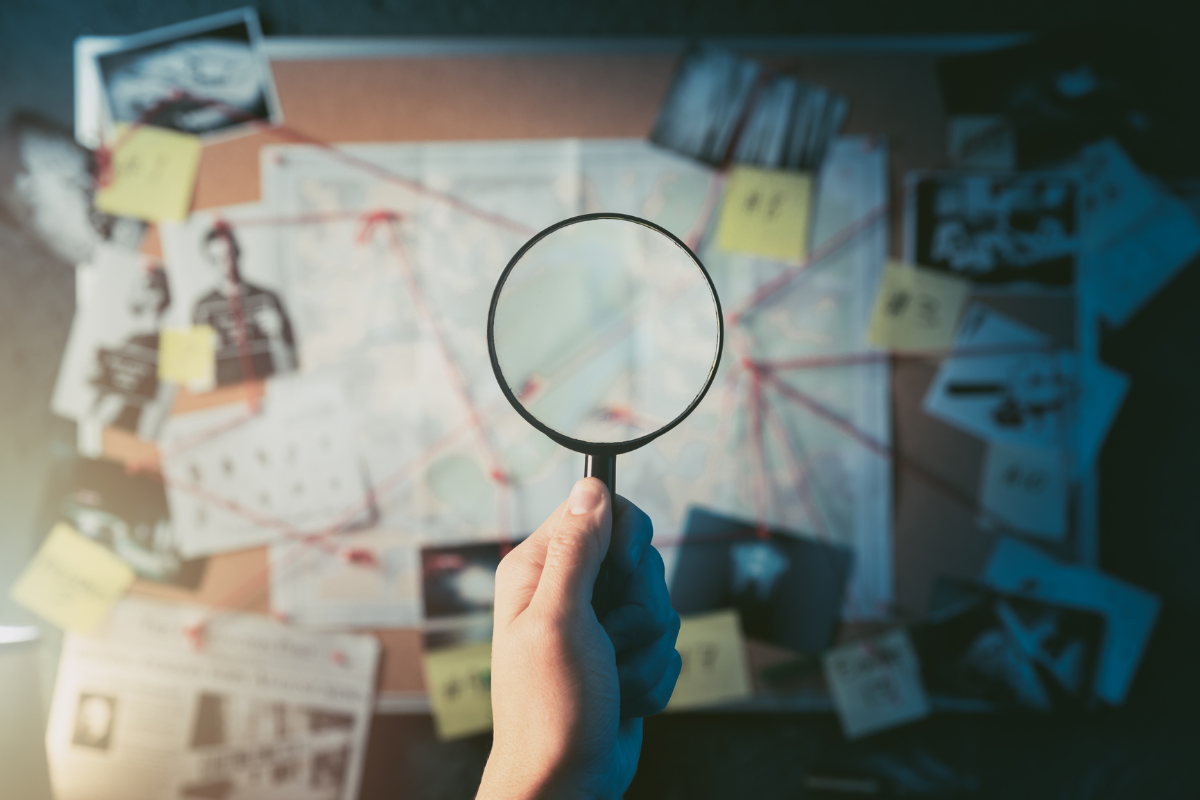 Hand holding magnifying glass up in front of a detective's evidence board.