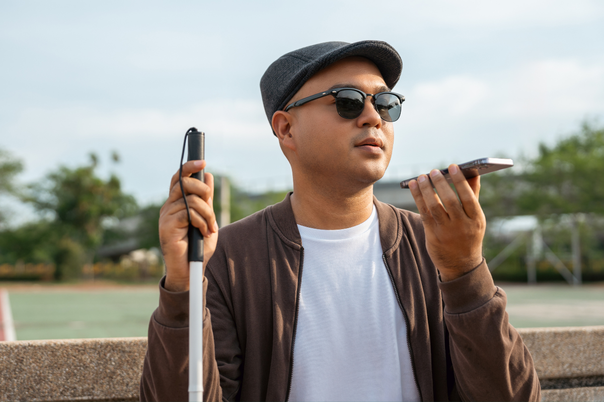 A man in sunglasses and a cap stands outside, holding a white cane and using a smartphone for directions.