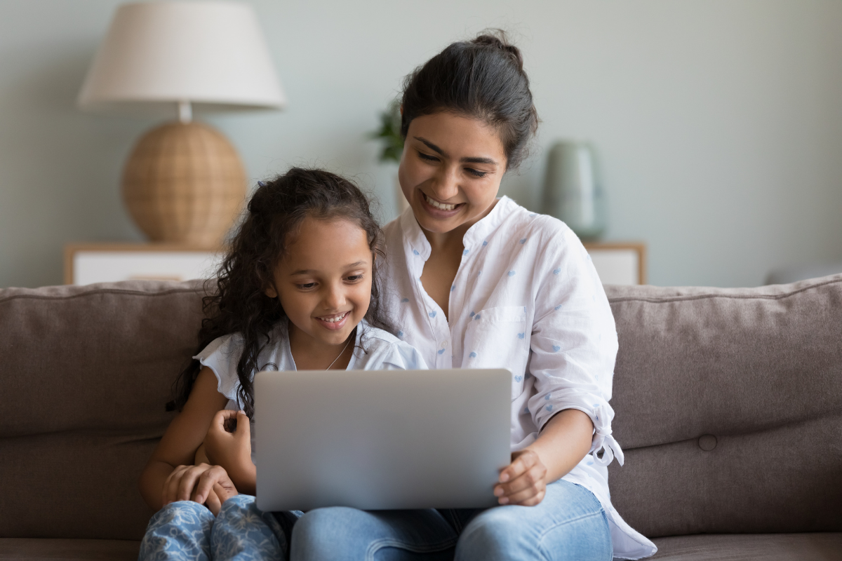 Smiling mother and young daughter sit on a couch, using a laptop together.